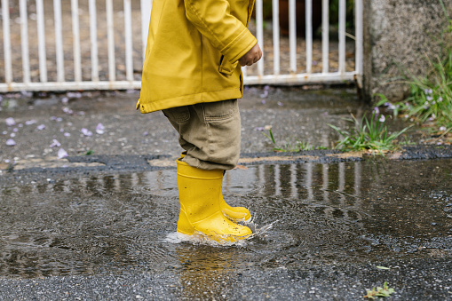 Cropped little playful kid in yellow raincoat and rubber boots walking in puddle and splashing water while having fun after rain. Warm clothing concept for winter and rainy days