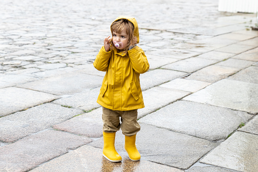 Full body of cute little kid wearing bright yellow raincoat and rubber boots standing on wet pavement and looking away. Concept anger, pissed off children and babies