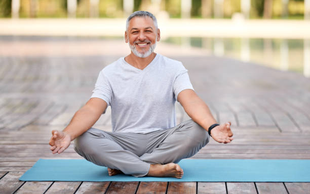 Calm happy senior man sitting in lotus pose on mat during morning meditation in park Calm happy senior man sitting in lotus pose on mat during morning meditation in park, holding hands in mudra gesture and smiling at camera. Mental health and yoga concept good posture photos stock pictures, royalty-free photos & images