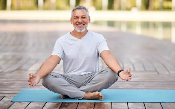 Calm happy senior man sitting in lotus pose on mat during morning meditation in park, holding hands in mudra gesture and smiling at camera. Mental health and yoga concept
