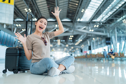 lockdown is over time to travel,happiness asian femlae casual cloth hand wave gesture smiling while sit relax on terminal airport floor with luggage safety travel concept