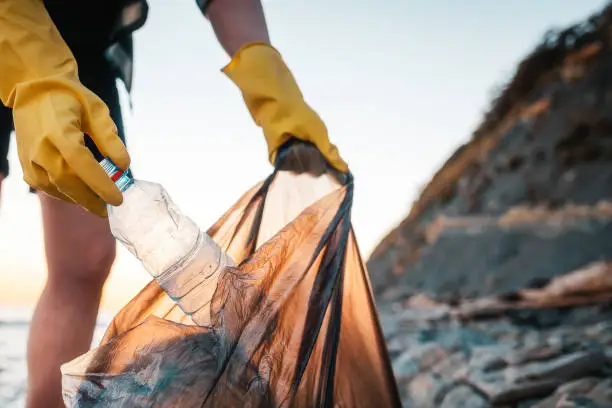 Photo of A woman volunteer puts a plastic bottle in a polyethylene bag. Close up of hands. In the background wild beach and ocean. The concept of environmental conservation and clean up of coast