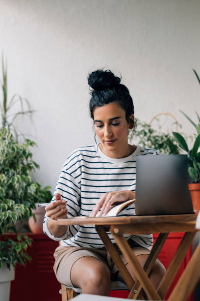 istock Beautiful Happy Woman Sitting Surrounded by Potted Plants, Writing Down Notes and Using her Laptop 1339854946