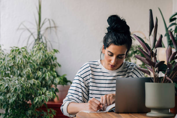 istock Beautiful Happy Woman Sitting Surrounded by Potted Plants, Writing Down Notes and Using her Laptop 1339854954