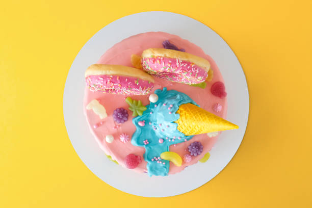 Cake on birthday with multicolored rainbow cream on a yellow background decorated with a waffle cone, pink donuts, sprinkles, marmalade candies poured with chocolate.