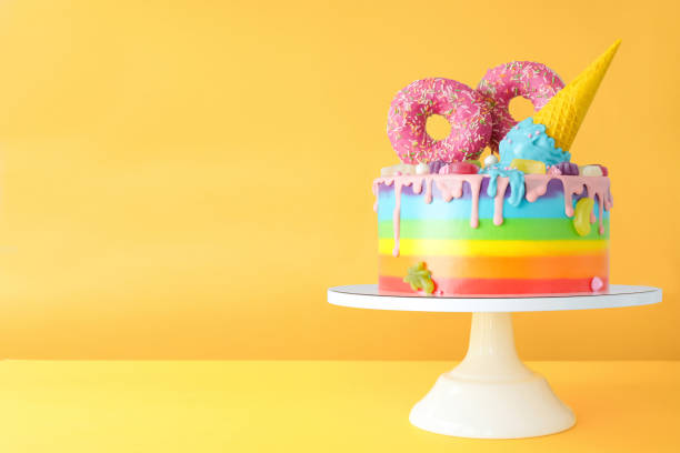 Cake on birthday with multicolored rainbow cream on a yellow background decorated with a waffle cone, pink donuts, sprinkles, marmalade candies poured with chocolate.