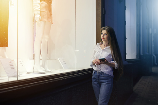 Lighted showcase of clothing store, near stands young woman with smartphone in his hands and looks inside on goods in evening time