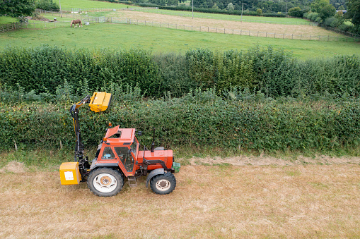 Drone point of view of an hydraulic tractor mounted hedge cutter being driven along the edge of an agricultural field. The farmer is cutting the high growth of the hedgerow, as part of his Autumn farmland maintenance.
