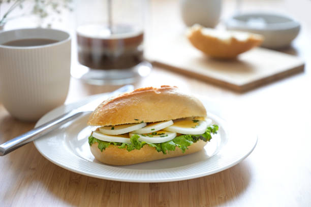 Sandwich roll with egg slices, lettuce and chives garnish on a white plate and fresh coffee on a light wooden breakfast table, selected focus, narrow depth of field