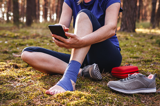 Injured hiker with sprain ankle using smart phone to get help. Woman gave herself first aid during hiking injury in nature