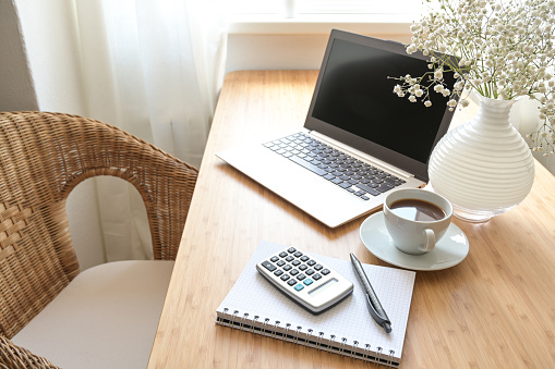 Home office desk with laptop computer, calculator, spiral book, coffee and a white flower bouquet, business accounting concept, copy space, selected focus