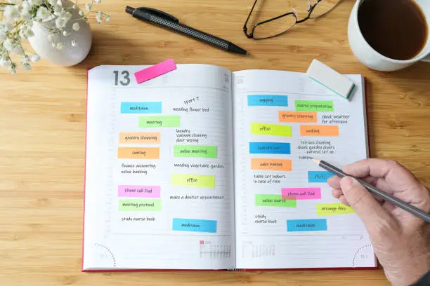 Hand of a woman writing appointments in a diary organizer or schedule calendar with a lot of colorful sticky notes for her time management on a wooden desk with coffee, flowers and glasses, selected focus, narrow depth of field