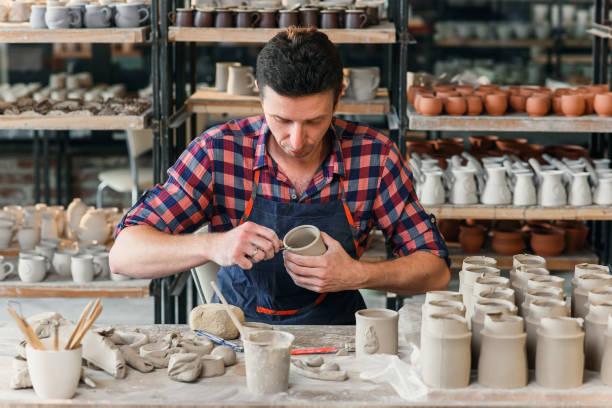 istock A middle-aged man in a work uniform make a clay shape of cup in a large creative pottery workshop. 1339882422