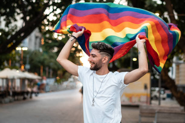 un jeune homme gay tient un drapeau arc-en-ciel - gay pride flag gay pride gay man homosexual photos et images de collection