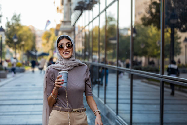 Mid adult woman with cup of coffee on city street in morning. Smiling Muslim woman wearing hijab headscarf walking in the city center. Arab female in urban background arab culture stock pictures, royalty-free photos & images