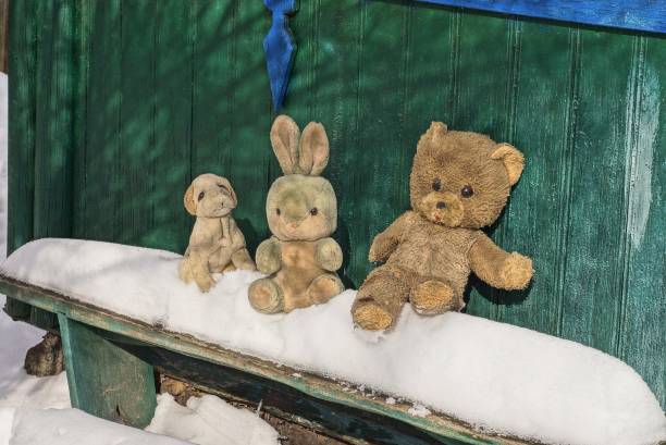 three old plush toy animals stand on a wooden bench in white snow near a green wall on a winter street