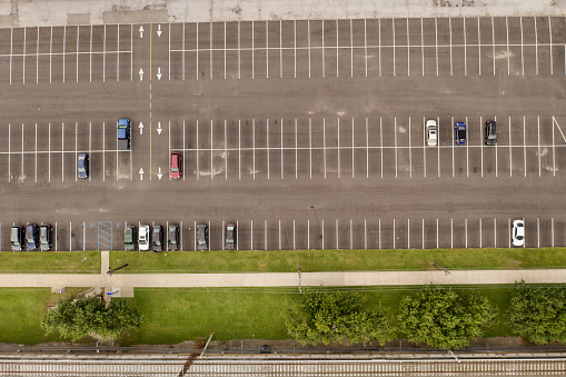 A top-down shot directly over an automobile parking lot with a few parked cars on a cloudy day on Long Island, NY. It is near a train station with a strip of grass on one side of the lot.