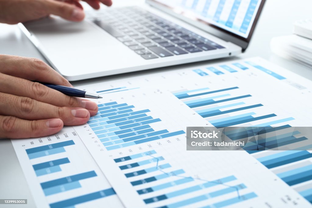 Analyzing business accounting. Man reviewing charts on reports and laptop display. Chart Stock Photo