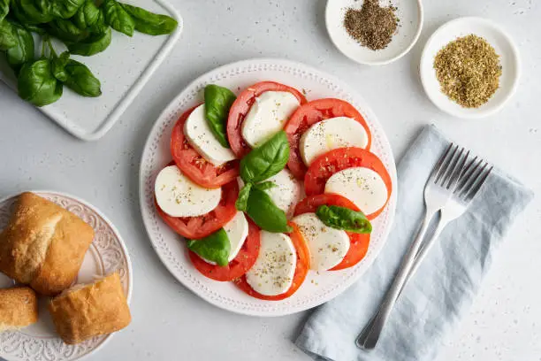 Delicious italian caprese salad with sliced tomatoes, mozzarella cheese, fresh basil, olive oil, pepper, salt and ciabatta. Mediterranean healthy food. Horizontal top view of appetizer