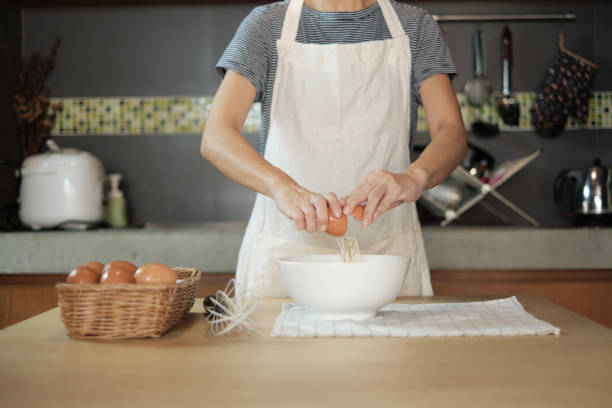 Female cook in a white apron is cracking an egg in home's kitchen.