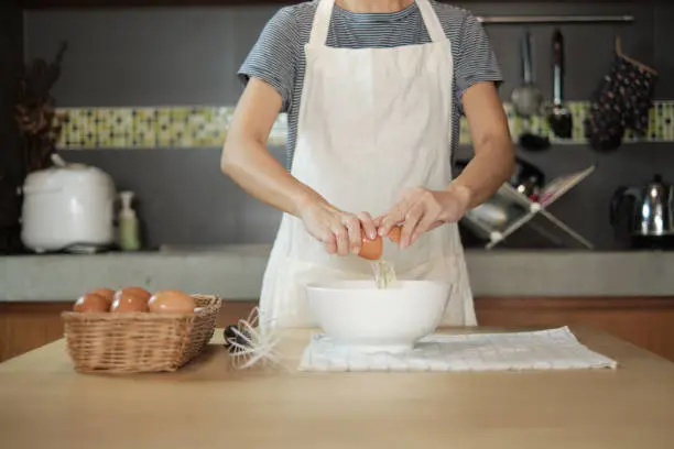 Close-up front view footage, a female cook in a white apron is cracking an egg into a cup to prepare a meal on a wooden table in the home's kitchen. Eating egg yolks is a healthy breakfast.