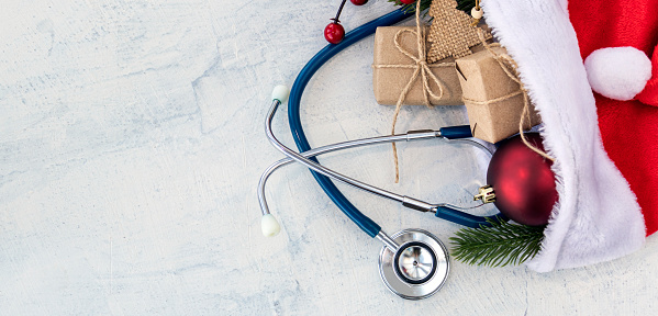 banner with Santa Claus hat and stethoscope, Christmas gifts, and a branch of Christmas tree with red Christmas ball on a white concrete table. Xmas. Copy space. Flat lay