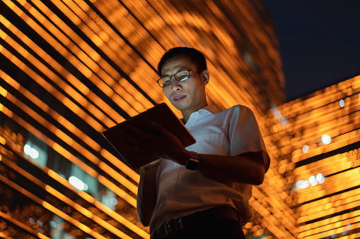An Asian male white-collar worker uses a tablet computer outdoors, with a light show on the exterior wall of an office building in the background