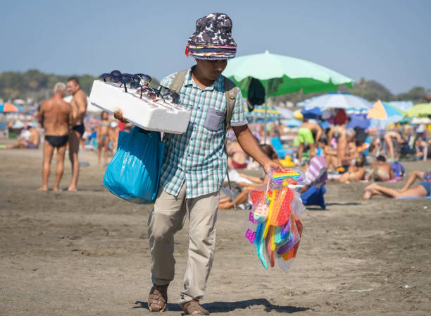 vendedor de sombreros en la playa entre multitudes de personas desenmascaradas en marina di san nicola, italia. - unmasked fotografías e imágenes de stock