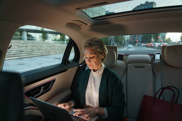 una donna d'affari dai capelli grigi in un'auto con il computer portatile. alleggerito dalla luce dello schermo del laptop - taxi travel business women foto e immagini stock