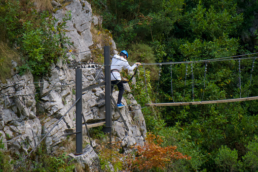 Back view of brave anonymous male hiker in protective helmet and harness walking on suspension bridge in mountains