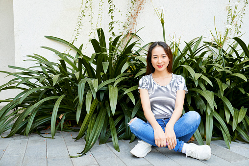 Cheerful beautiful young Vietnamese woman sitting on ground with her legs crossed and smiling at camera