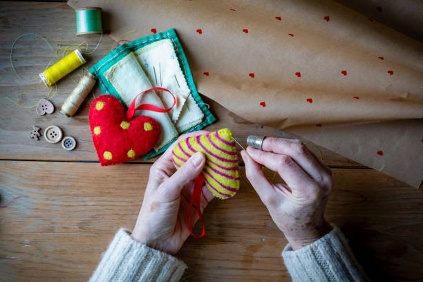 A table top view of an unrecognisable person sewing a homemade Christmas tree decoration on a wooden table top surrounded by wrapping paper, reels of thread, wooden buttons.