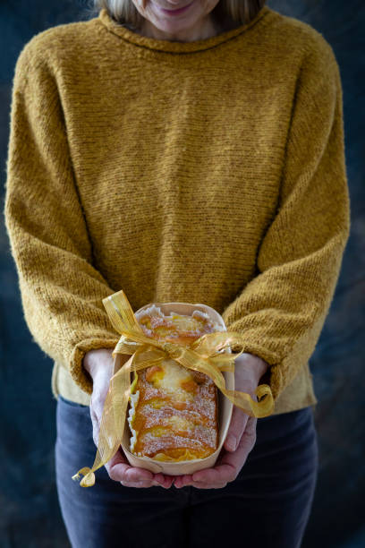 A mature caucasian woman holding a baked lemon cake in a reusable thin wooden box, decorated with a ribbon bow. This is a sustainable Christmas gift handmade at home.