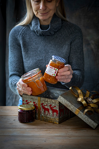 A caucasian senior woman filling a Christmas themed metal tin with homemade jam, marmalade and chutney in reused jars. These have been made as sustainable Christmas gifts.