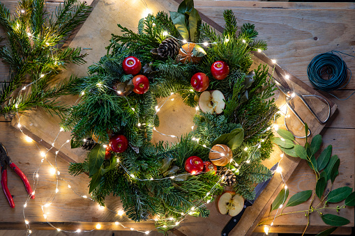A table top view of a freshly made homemade Christmas wreath on a wooden board with fairy lights draped over it. It is decorated with apples, pine cone, oranges and eucalyptus leaves and is ready to be given as a gift.