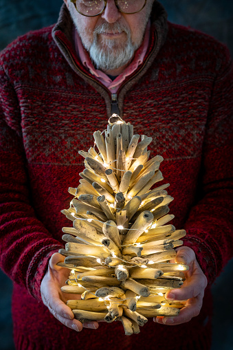 An unrecognisable man holding a homemade wooden Christmas tree with fairy lights on and showing it to the camera. It is illuminated and is going to be given as a gift at Christmas time.