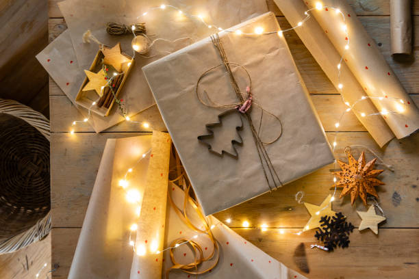 A table top view of a Christmas gift wrapped for Christmas on a wooden table top. It is decorated with string and a cookie cutter. On the table there is other decorations and items for wrapping such as paper, Christmas decorations, string and the table is draped with Christmas lights.