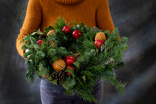 An unrecognisable woman holding a freshly homemade Christmas wreath and showing it to the camera. It is decorated with apples, pine cones, oranges and eucalyptus leaves and is ready to be given as a gift.