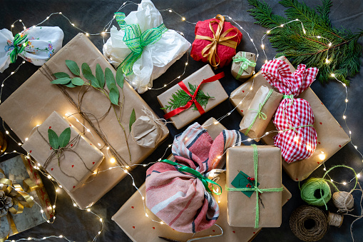 An overhead view of wrapped Christmas presents on a backdrop which have been decorated with ribbon, string, eucalyptus leaves and holly leaves. They are ready to be given at Christmas time and they are on the table, draped in Christmas lights.
