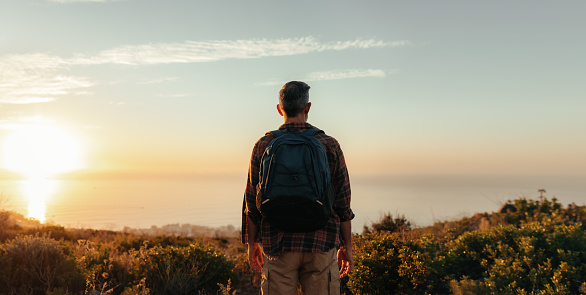 Rearview of a backpacker looking at the panoramic view on a hilltop. Unrecognisable male hiker standing alone on a coastal hill. Adventurous mature man enjoying the sunset outdoors.
