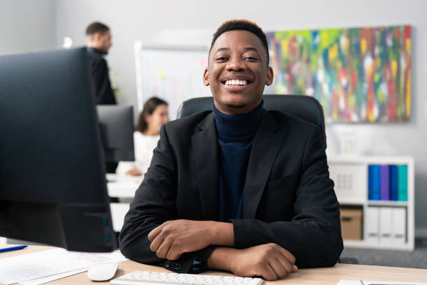 Sympathetic smiling new corporate office worker, male wearing carny jacket and turtleneck, looks into camera with big brown eyes, white teeth, dark skin, working behind desk in front of computer