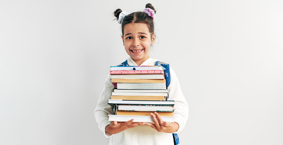 Smiling little girl wearing uniform, backpack holding pile of books on her arms. Schoolgirl carrying lot of books while going to school against grey studio background.