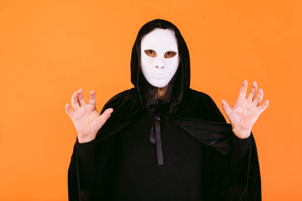 Portrait of a person with white Halloween killer mask, cape and hood looking at camera, scaring with hands, dressed for Halloween over orange background stock photo