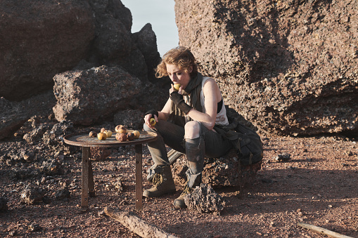 Homeless young woman sitting on the rock and eating potatoes outdoors