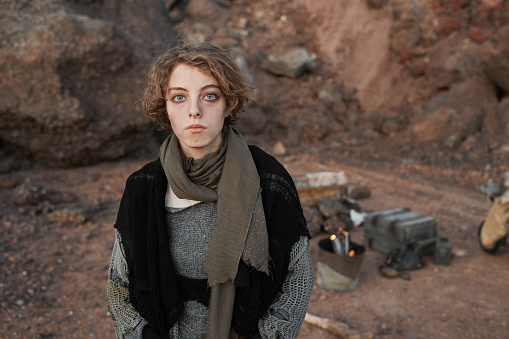 Portrait of young homeless woman in torn clothes looking at camera while standing outdoors