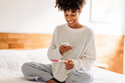 Woman taking a picture of her home pregnancy test. Excited young woman capturing her pregnancy test results with a smartphone. Woman smiling cheerfully while sitting on her bed at home.