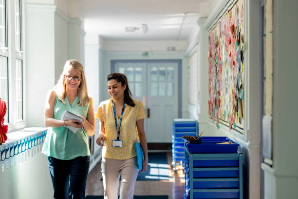 Sharing Teaching Ideas A front view of two female teachers sharing notes and teaching ideas as they walk along the corridor of the school they work at in Hexham in the North East of England. They are smiling and laughing as they walk along to their classrooms. school building stock pictures, royalty-free photos & images