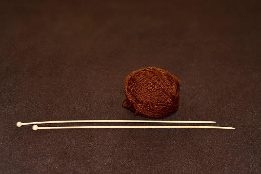 A ball of brown woollen threads and two wooden spokes close-up. Preparation for knitting. Brown background.
