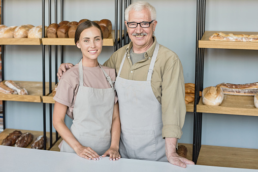 Portrait of happy senior baker in apron embracing young daughter while working with her in own bakery shop