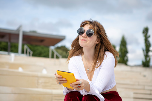 Fashionable student with an e-book outside in summer - she is wearing a white top, red velvet flared trousers and sunglasses
